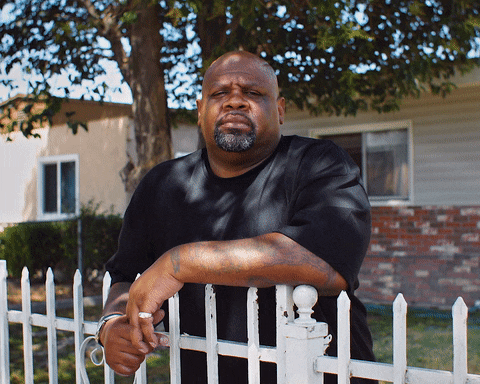 Cinemagraph of Facing Life subject Melvin Smith leaning on the fence in front of his house. The tree in the background and show it casts are moving.