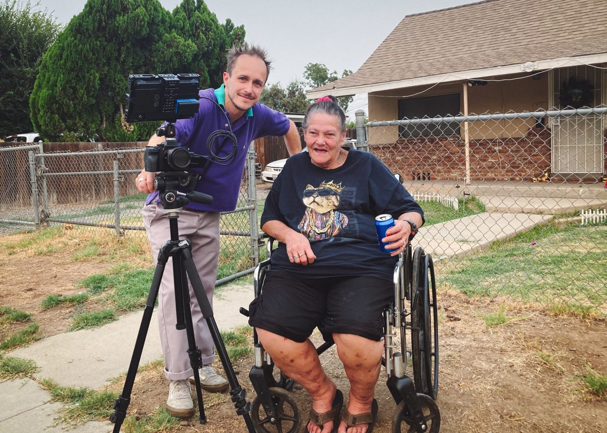 Tauszik (left) stands with a video camera next to Facing Life subject Robin Marlowe who is in  a wheelchair and holding a blue can.