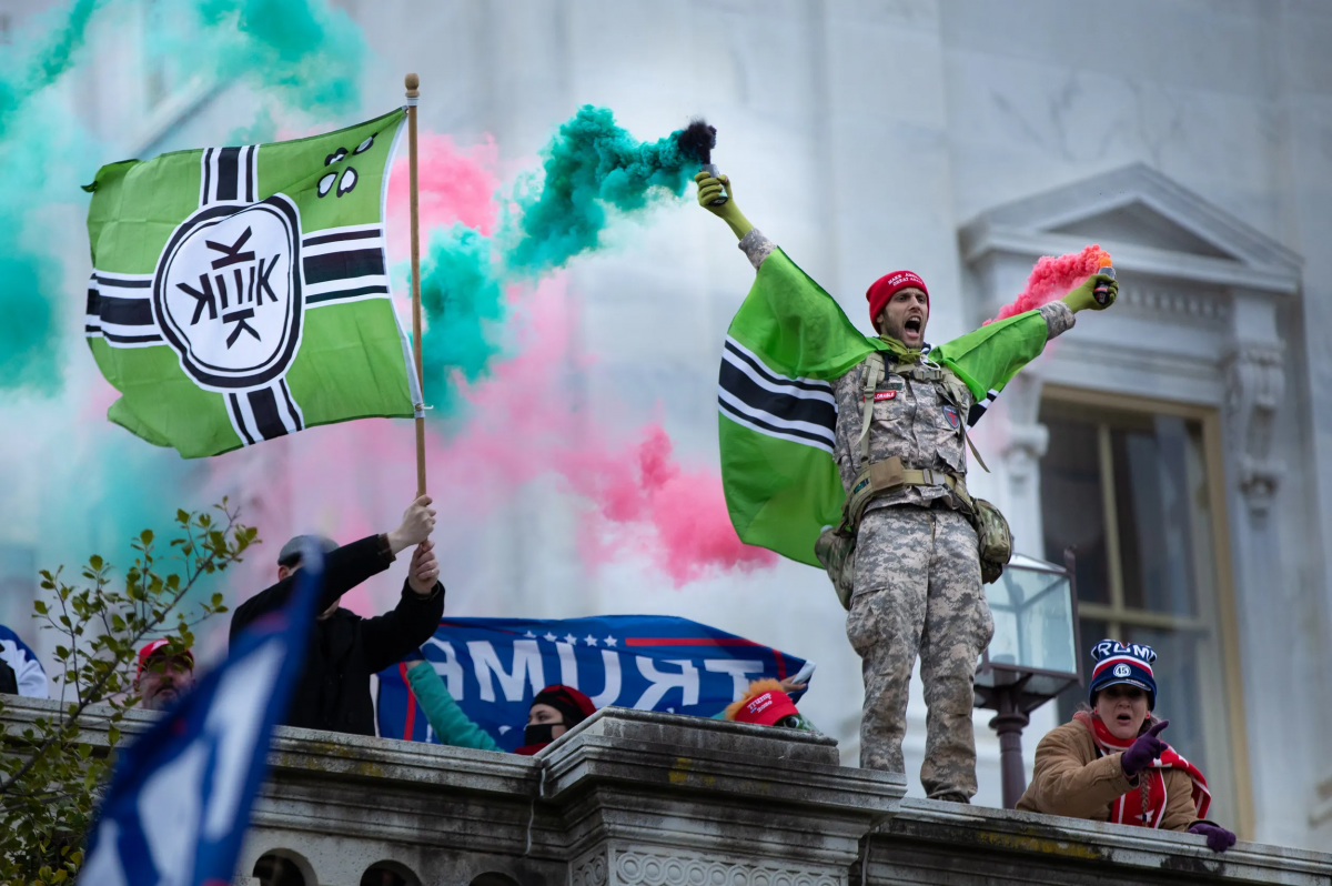Image of an alt-right man wearing military fatigues and wearing a red beanie standing at the capital with green and pink smoke bombs in his hands. On his shoulders, and being waved by another man to the left, is the Kekistan flag. The flag is green with a black and white cross. In the center is a circle with four Ks. Behind them a Trump flag is being held.