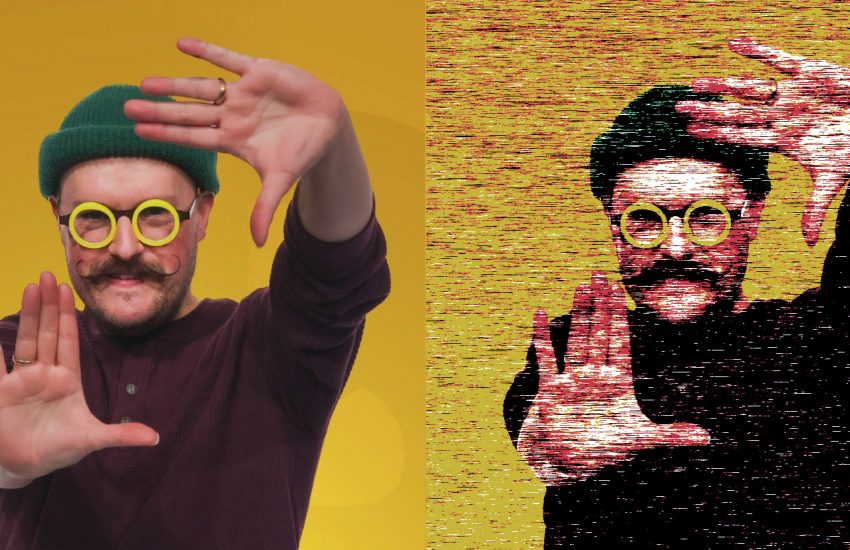 Man making framing gesture against a yellow background. He's wearing round yellow glasses and a black beanie.